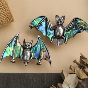 Może przedstawiać: Dwie srebrne broszki w kształcie nietoperzy z iryzującymi skrzydłami. Skrzydła prezentują żywą mieszankę odcieni zieleni, błękitu i fioletu. Nietoperze są szczegółowe z małymi uszami i teksturowanym korpusem. Broszki są prezentowane na neutralnym tle.