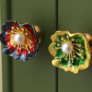 Peut inclure: Deux boutons de meuble décoratifs en forme de fleur. L'un est rouge, jaune et bleu avec un centre de perle et des accents de strass. L'autre est vert et or avec un centre de perle et des accents de strass. Les deux ont une quincaillerie en laiton.