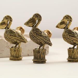 Puede incluir: Tres tiradores de armario de latón con forma de pelícano. Cada tirador presenta una figura detallada de un pelícano posado sobre un poste de madera. Los tiradores tienen un aspecto vintage y antiguo, con un tono dorado cálido. Los pelícanos miran en diferentes direcciones.