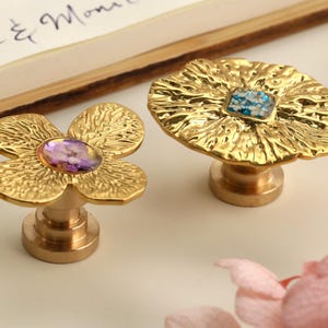 Peut inclure: Deux boutons de meuble décoratifs dorés. L'un est en forme de fleur avec un centre violet, et l'autre est de forme ronde avec un centre bleu et blanc. Les deux boutons ont un design texturé et sont sur une surface blanche.