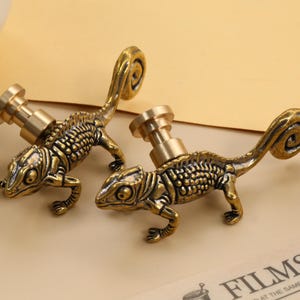 Pode incluir: Dois puxadores de armário de camaleão de latão antigo. Os puxadores apresentam um design detalhado de lagarto com um poste de parafuso de latão. Os lagartos estão em uma pose de caminhada com a cauda enrolada. Ferragens decorativas.