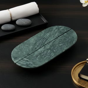 Könnte beinhalten: Ein grünes ovales Tablett aus Marmor mit geteiltem Design, neben einem schwarzen Tablett mit einem gerollten weißen Tuch und zwei grauen Steinen. Eine kleine Vase mit weißen Blumen und ein goldenes Tablett mit einem schwarzen Notizbuch und Stift sind ebenfalls vorhanden.