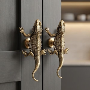 May include: Two brass lizard-shaped cabinet handles mounted on a gray cabinet door. The handles are detailed with textured scales and outstretched limbs. The cabinet door is part of a kitchen setting.
