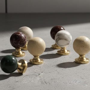 Könnte beinhalten: Eine Sammlung dekorativer Schrankknöpfe. Die Knöpfe haben kugelförmige Oberteile in verschiedenen Farben und Mustern, darunter grüner, brauner, beiger und weißer Marmor. Jeder Knopf ist auf einem goldfarbenen Sockel montiert.