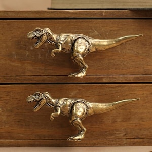 Könnte beinhalten: Eine Aufbewahrungseinheit aus Holz mit zwei Schubladen, die jeweils mit einer metallisch goldfarbenen Tyrannosaurus Rex-Dinosaurierfigur als Griff versehen sind. Die Dinosaurierfiguren sind horizontal nach links ausgerichtet und detailliert.
