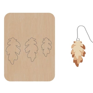 Puede incluir: Una hoja de madera con tres recortes de hojas de roble y un solo pendiente de hoja de roble con un degradado de color marrón, beige y crema.