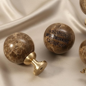 Op de afbeelding: Marmeren bolvormige kastknoppen met messing voetjes. Op één knop staat het woord "WOODWORKING" gegraveerd. De knoppen zijn bruin met tan adering. De knoppen liggen op een crèmekleurig satijnen oppervlak.