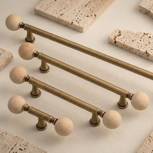 Könnte beinhalten: Vier Schrankgriffe mit Messing-Finish und kugelförmigen, hellfarbigen Holzknöpfen. Die Griffe sind in einer Reihe angeordnet, mit unterschiedlichen Längen. Beige Steinplatten sind um die Griffe herum verstreut.