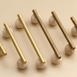 Könnte beinhalten: Eine Sammlung von Messing-Schrankgriffen, jeweils ein gerader Stab, ruht auf kleinen, runden, strukturierten Steinkugeln. Die Griffe variieren leicht in der Farbe, von hellem Gold bis zu dunkler Bronze. Der Hintergrund ist einfarbig beige.