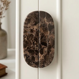 May include: A pair of semi-circular cabinet pulls made of dark brown marble with white veining. The two pieces are positioned together to form a single oval shape, mounted vertically on a white cabinet door with a smooth, polished finish.