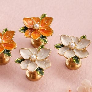 May include: Four decorative flower-shaped cabinet knobs. Two are orange, and two are white, each with a pearl center and green leaves. The knobs have a gold-colored base and are set against a pink background.