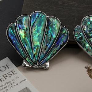 Peut inclure: Deux objets décoratifs en forme de coquillage aux tons irisés verts, bleus et argentés. Les coquillages ont une surface texturée et détaillée et sont posés sur un fond neutre. Il s'agit probablement de bijoux ou d'accessoires décoratifs.