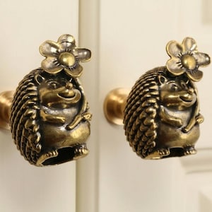 May include: Two antique brass hedgehog cabinet knobs with flower accents. The knobs are detailed with a smiling hedgehog design and a flower on top. The knobs are mounted on a white cabinet door.
