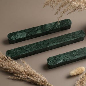 May include: Three rectangular bars made of dark green marble with natural veining. Each bar features rounded ends and a smooth, polished surface. The bars are arranged diagonally across the frame, showcasing the deep green stone texture.