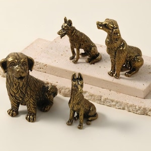 May include: Four bronze dog figurines in different poses are displayed on a layered, textured, off-white surface. The dogs include a Labrador, a German Shepherd, and a small puppy. The figurines have detailed features and a metallic finish.