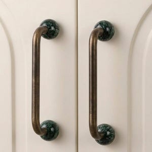 May include: Two vertical cabinet handles featuring a long, brushed bronze-toned metal bar connected to two spherical dark green marble mounts. The handles are mounted on a pair of off-white cabinet doors with arched panel detailing.