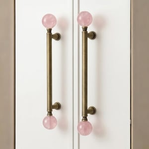 May include: Two vertical cabinet handles featuring a cylindrical antique brass finish bar with a spherical light pink stone knob at both the top and bottom ends. The hardware is mounted onto a white cabinet door with a central vertical seam.