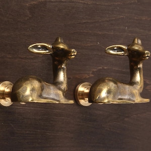 Puede incluir: Dos tiradores de armario con forma de ciervo de latón. Cada tirador presenta un ciervo reclinado con cuernos y orejas detallados. Los tiradores están montados sobre una base circular, de metal dorado.