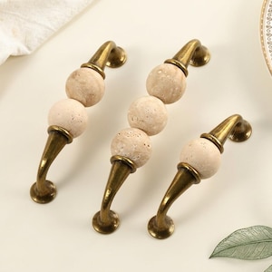 May include: Three cabinet pulls featuring antique brass metal hardware with curved ends and spherical, porous beige stone accents. Two of the pulls have two stone spheres, while the third pull features a single stone sphere in the center.