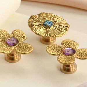May include: Gold-colored flower-shaped cabinet knobs with textured petals and a central gemstone. The knobs have a brass base and are arranged on a neutral surface. The gemstones are purple and blue.