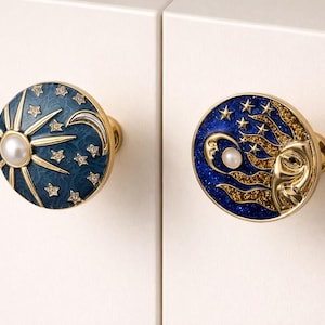 Pode incluir: Dois puxadores de armário com tema celestial. Um puxador apresenta um sol e uma pérola, o outro uma lua e estrelas, tudo sobre um fundo azul escuro. Os puxadores têm ferragens douradas e são adornados com pequenos cristais.