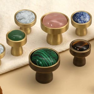 May include: A collection of round cabinet knobs featuring brushed brass bases and inlaid circular stone tops. The stones include white marble, green aventurine, pink quartz, blue sodalite, orange carnelian, tiger's eye, and green malachite patterns.