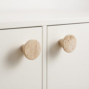Può includere: Primo piano di due pomelli per armadietti in pietra beige attaccati a un armadio bianco. I pomelli sono rotondi con una faccia piatta e una base cilindrica. La pietra ha una consistenza macchiettata. Le ante dell'armadio sono a filo e verniciate di bianco.