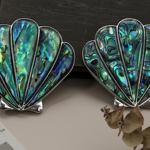 May include: Two shell-shaped ornaments with iridescent green, blue, and silver hues. The shells have a detailed, textured surface and are set against a neutral background. These are likely jewellery or decorative items.