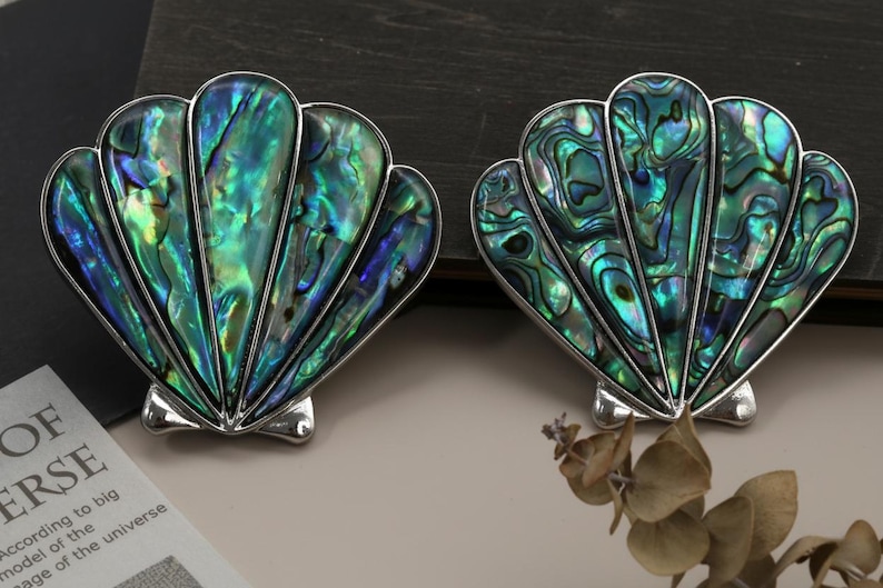 May include: Two shell-shaped ornaments with iridescent green, blue, and silver hues. The shells have a detailed, textured surface and are set against a neutral background. These are likely jewellery or decorative items.