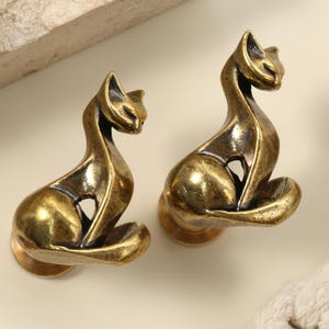 Könnte beinhalten: Zwei antike, messingfarbene Schrankknöpfe in Katzenform. Die Knöpfe haben ein stilisiertes Design einer sitzenden Katze mit gebogenem Schwanz und kleinem kreisförmigen Sockel. Das Messing hat eine warme, gealterte Patina.