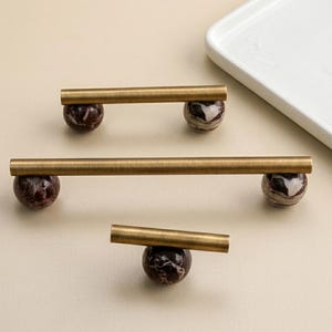 May include: Three brass cabinet pulls with dark red and brown marble sphere accents. The pulls are arranged on a light beige surface, with a white rectangular tray in the background. The hardware is designed for furniture.