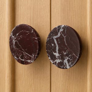 May include: Two circular cabinet knobs made of dark burgundy marble with prominent white veining. The knobs are mounted on a light-colored wooden surface with vertical paneling, showcasing a smooth, polished finish and a rounded, flat-faced design.