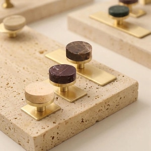 May include: A collection of circular cabinet knobs featuring natural stone tops in shades of beige, dark brown, and deep purple, each mounted on a brushed brass rectangular base. The knobs are displayed on a textured, porous beige stone slab.