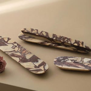 May include: Three elongated oval trays crafted from polished marble featuring a distinct brecciated pattern with deep burgundy and cream-coloured veining. The set includes two long, narrow trays and one smaller, compact oval tray, accompanied by a dried rose.