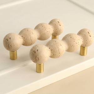 May include: Two cabinet pulls featuring a series of connected, spherical beige stone beads with a porous, natural texture. Each handle is supported by two cylindrical, metallic gold-toned posts that attach to the base of the stone structure.