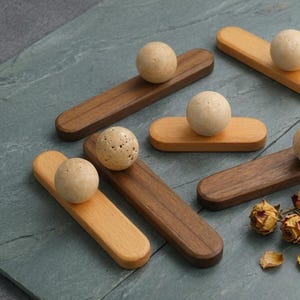 May include: Wooden chopstick rests in various shades of brown and tan, each featuring a spherical stone accent. The rests are arranged on a textured, gray-green surface, with dried flowers in the corner.