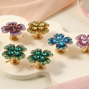Puede incluir: Un conjunto de tiradores decorativos para armarios con forma de tréboles de cuatro hojas. Cada tirador presenta cristales brillantes de diferentes colores, como morado, amarillo, azul y rosa, sobre una base dorada. Los tiradores están dispuestos sobre una superficie blanca.