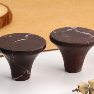 Puede incluir: Un juego de pomos para armarios de mármol marrón oscuro con un acabado liso y pulido y vetas blancas distintivas. Cada pomo presenta una parte superior circular ancha y plana que se estrecha hacia una base cilíndrica, creando una forma.