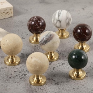 Op de afbeelding: Een verzameling decoratieve kastknoppen met bolvormige stenen toppen in verschillende kleuren en patronen, waaronder beige, bruin, wit en groen. Elke knop is gemonteerd op een messing voet, wat een klassieke en elegante uitstraling biedt.