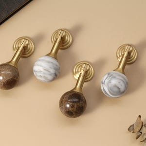 May include: Four cabinet knobs with brass hardware and marble spheres. Two knobs have brown marble spheres, and two have white and gray marble spheres. The knobs are arranged on a light beige surface.
