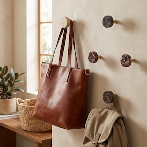 Puede incluir: Un bolso tote de cuero marrón cuelga de un gancho de pared de madera. Ganchos adicionales de mármol y latón están montados en la pared. Una mochila de lona color beige está suspendida de un gancho de mármol. Se ve una cesta tejida y una planta en maceta.