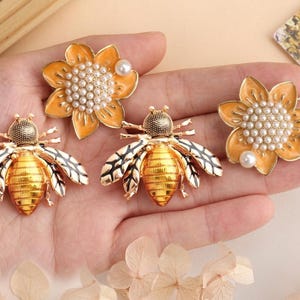 May include: A selection of decorative brooches with bee and flower designs. The bee brooches have gold-coloured bodies and black and white wings. The flower brooches are orange with pearl-like centres and a single pearl accent.
