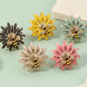Peut inclure: Cinq broches en forme de fleur de différentes couleurs : gris, jaune, turquoise, rose et beige. Chaque broche présente une abeille détaillée au centre. Les broches sont posées sur un fond vert clair avec des accents de pierre blanche.