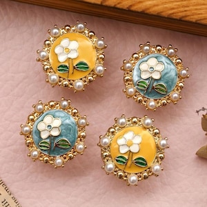 May include: Four floral stud earrings. Each earring features a white flower with green leaves on a yellow or teal background, surrounded by a gold-coloured frame and small pearls. The earrings are set on a pink surface.