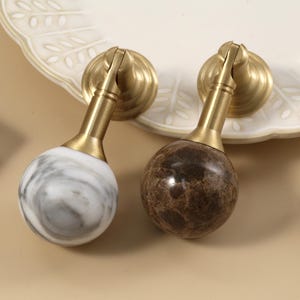 Peut inclure: Deux poignées de meubles en laiton avec des sphères en marbre, une blanche et grise, l'autre marron et beige. Les poignées ont un design classique, avec une base ronde et une poignée incurvée. Une troisième poignée est visible à droite.