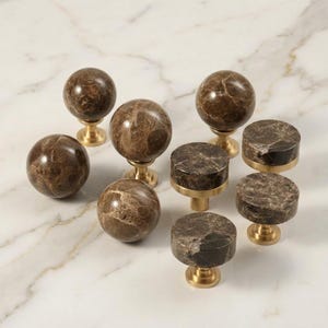 Peut inclure: Ensemble de boutons et poignées d'armoire en marbre brun. Les boutons sont sphériques et cylindriques, avec des bases en laiton. Le marbre présente un motif veiné naturel. La quincaillerie est disposée sur une surface en marbre blanc.