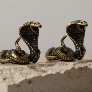 May include: Close-up of three bronze cobra figurines with open mouths, coiled bodies, and detailed scales. The statues are on a textured, light-colored surface, with a neutral background. The cobra statues are a decorative item.