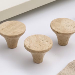 Peut inclure: Ensemble de trois boutons de meuble en pierre beige, avec un dessus arrondi et une base évasée. Les boutons sont en pierre tachetée et sont conçus pour les tiroirs ou les armoires. Les boutons sont présentés sur une surface blanche.