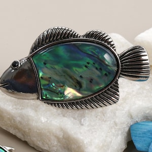 Peut inclure: Broche argentée en forme de poisson avec un centre en coquillage d'ormeau irisé vert et bleu. La broche présente des nageoires texturées et un petit œil noir. Elle est posée sur une pierre blanche.