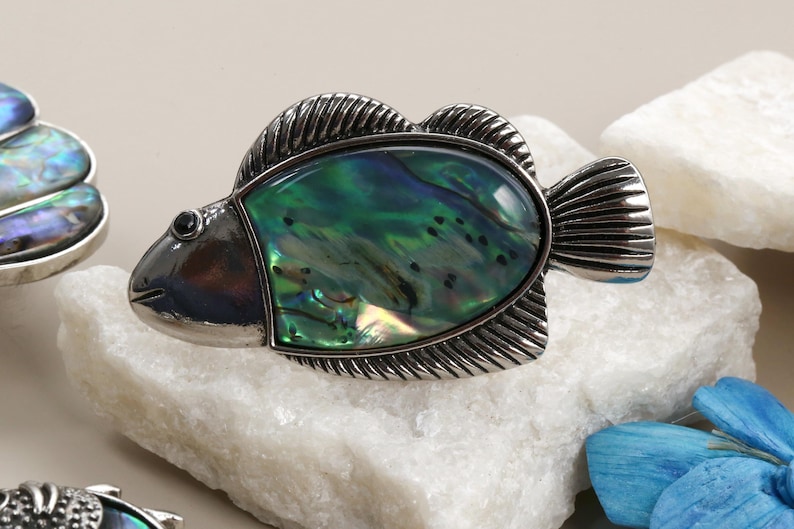 May include: A silver fish brooch featuring an iridescent green and blue abalone shell. The brooch has textured fins and a small black eye. It is displayed on a white stone. The brooch is a decorative accessory.